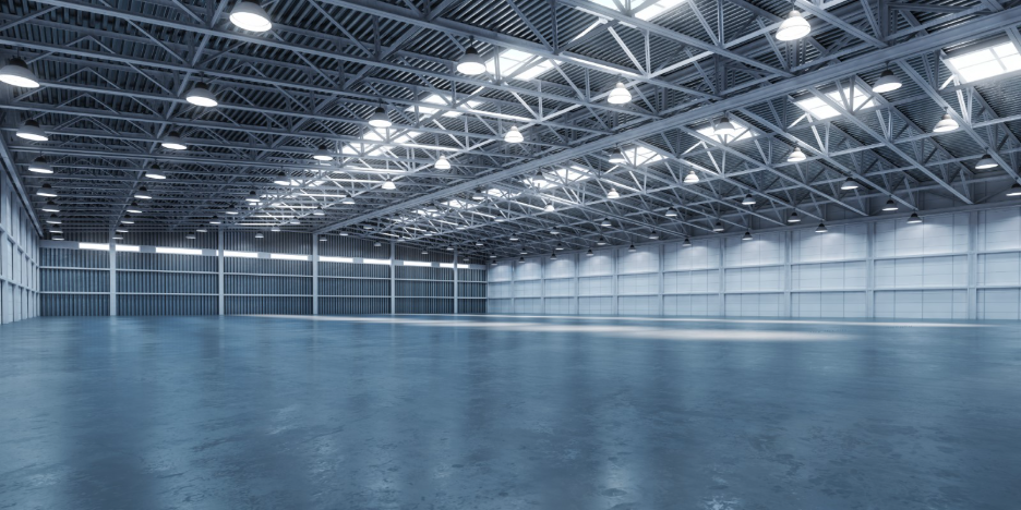 A large, empty industrial warehouse with a spacious polished concrete floor, exposed steel roof trusses, numerous overhead lights, skylights, and corrugated metal walls, emphasizing the building’s open interior and expansive scale.